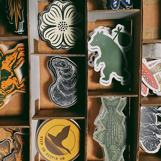 A collection of various stickers organized in a cardboard box. The stickers include nature designs such as animals, flowers, and abstract patterns.