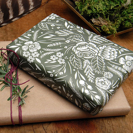 Two wrapped gifts are shown. One is covered in green floral wrapping paper, and the other is wrapped in plain brown paper with a sprig of herbs tied with string. Both rest on a wooden surface.