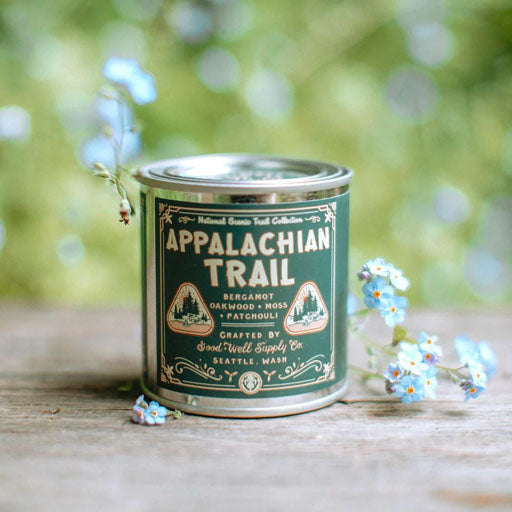 A small green tin candle labeled "Appalachian Trail" from Good & Well Supply Co. is placed on a wooden surface with light blue flowers in the background. The candle is scented with bergamot, oakmoss, and patchouli.