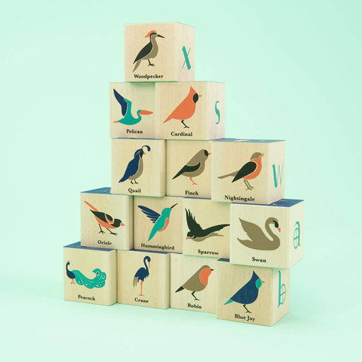 A pyramid of wooden blocks, each featuring an illustrated bird and corresponding bird name, including Woodpecker, Pelican, Cardinal, Quail, and others. The background is a solid light green.