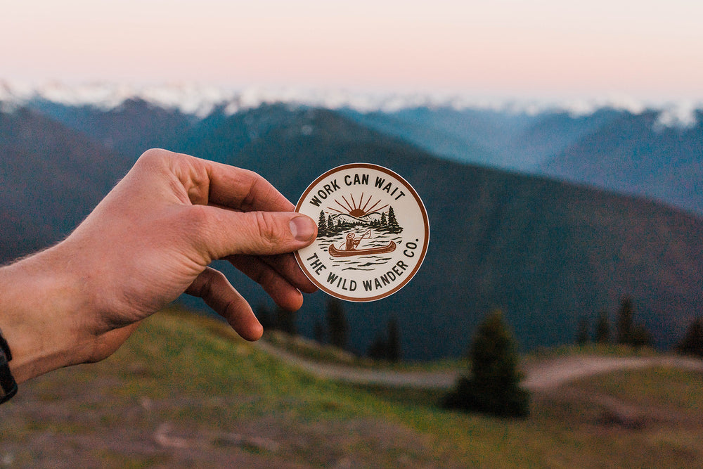 A hand holding a circular sticker with the text "WORK CAN WAIT" and "THE WILD WANDER CO." against a mountainous landscape at sunset.