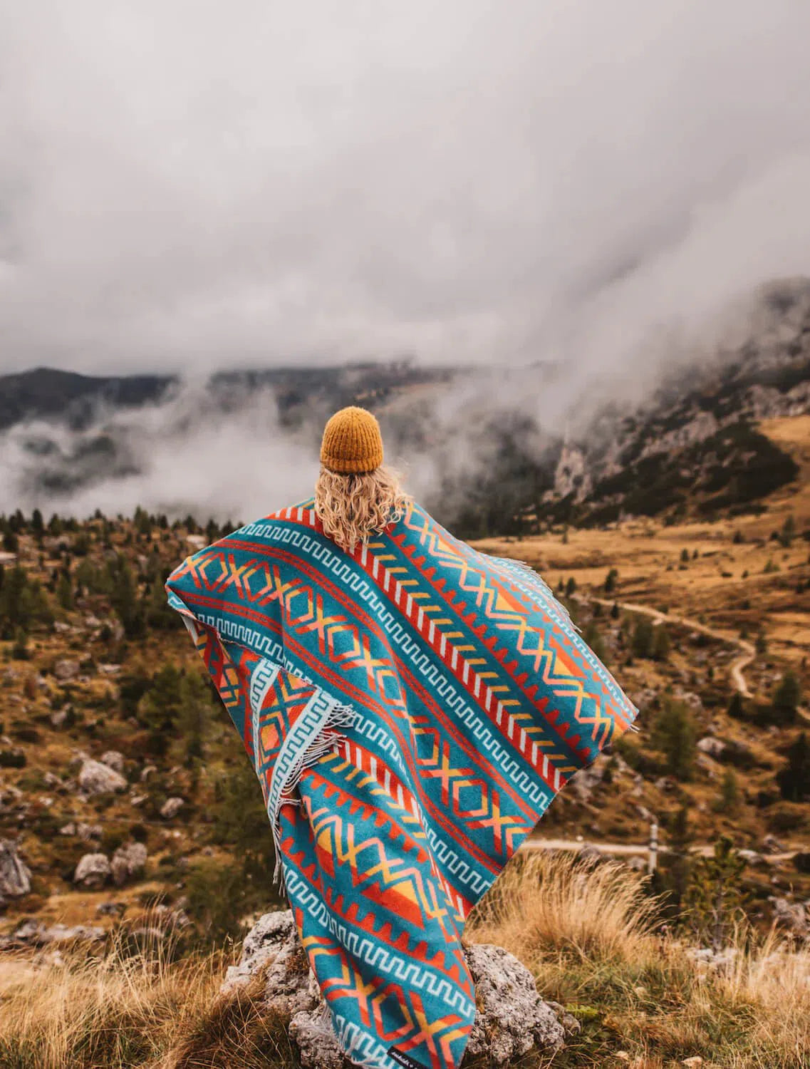 A woman wearing a colorful Ridgeline Sierras blanket on top of a mountain.